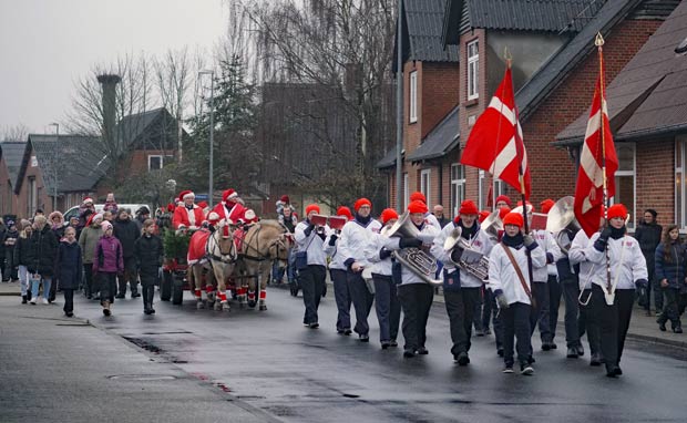 FDF-orkesteret anfører juleoptogets festlige parade, fulgt tæt af en hestevogn og en strøm af glade børn og voksne i nissehuer, der fejrer sæsonens glæder gennem byens gader